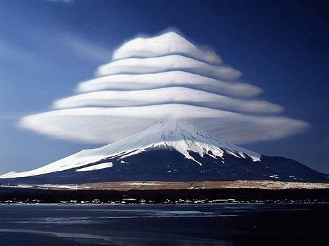 Extraordinary lenticular clouds stack in horizontal layers above a snow-capped volcanic peak, creating an almost surreal natural formation. A frozen lake stretches across the foreground, reflecting the deep azure sky.