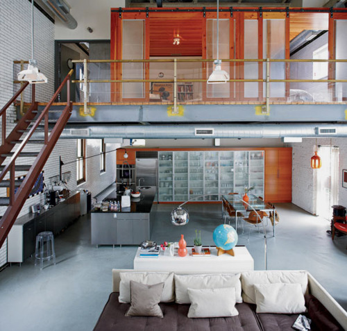 A two-story loft showcases polished concrete floors and metal railings beneath soaring ceilings with a mezzanine level above. The white modular sofa grounds the open living area while warm orange lighting from the upper level adds contrast to the industrial palette.