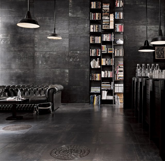 Floor-to-ceiling bookshelves line raw concrete walls creating a dramatic reading room with a leather Chesterfield sofa. Sculptural pendant lights illuminate the moody space carved from industrial architecture.