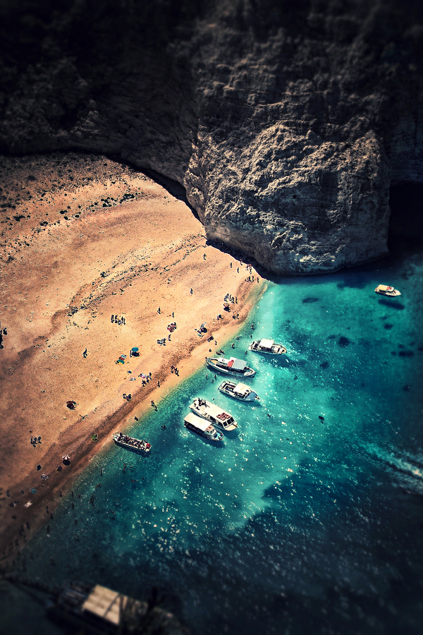 An aerial view reveals a secluded Mediterranean cove where golden sand meets turquoise water, surrounded by dramatic limestone cliffs. Small boats dot the crystalline bay while beachgoers enjoy the pristine shoreline.