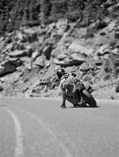 A motorcyclist leans into a sharp curve on an empty mountain road in this black and white composition, with rocky cliffs and scattered boulders creating dramatic vertical elements. The contrast between smooth asphalt and raw stone emphasizes human determination against natural terrain.