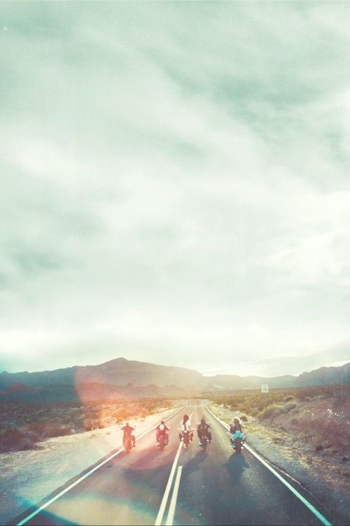 Motorcyclists stand in silhouette on an empty desert highway, backlit by brilliant sun lens flare beneath a cloud-filled sky transitioning from pale mint to warm amber. The dramatic mountain ranges and vintage aesthetic capture the romance of the open road.