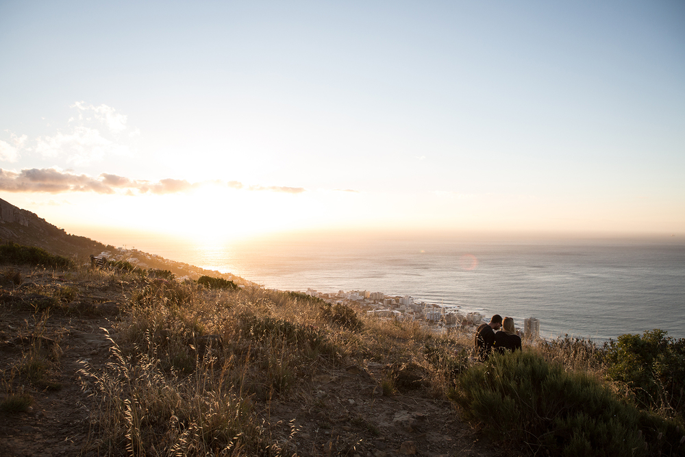 Two figures sit together on a scrub-covered coastal hillside overlooking the ocean during golden hour, surrounded by warm amber light. The rolling hills descend to the water's edge in layers of peachy and sage tones.