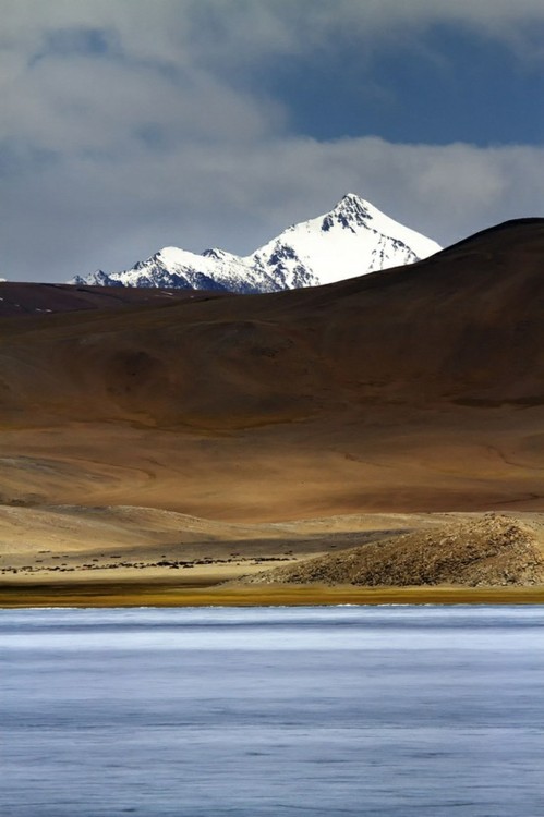 A high-altitude lake mirrors the sky beneath snow-capped peaks, while rolling desert hills create layered earth tones in the foreground. The remote landscape shows where mountain and desert ecosystems meet.