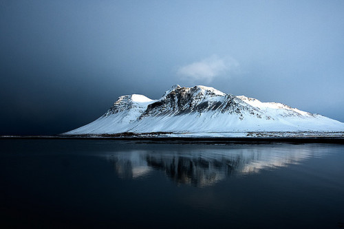 A snow-capped mountain rises from still waters under twilight, its peak perfectly mirrored in the glassy surface. The scene captures Nordic landscape geometry in near-perfect symmetry.