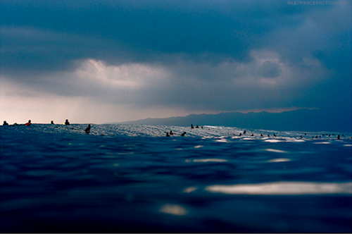 Surfers appear as silhouettes against rolling waves in this water-level perspective, with dramatic storm clouds meeting turbulent ocean swells along the horizon line.