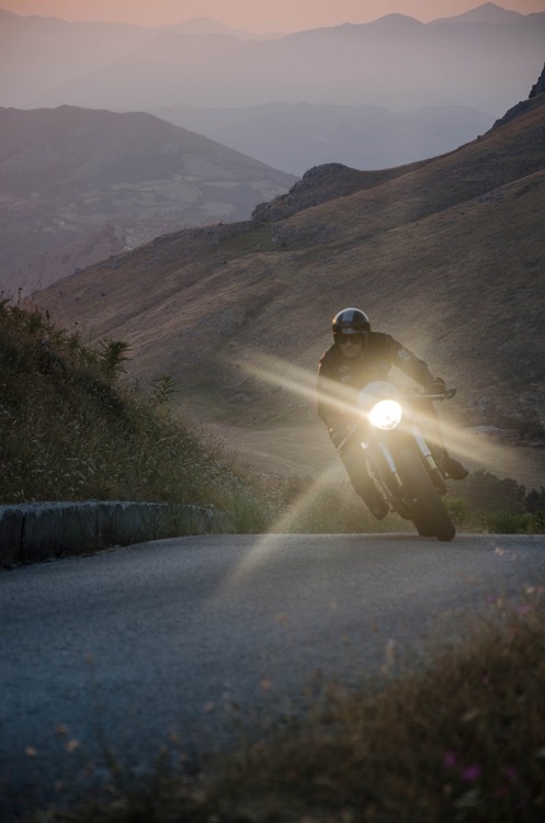A motorcyclist's headlight cuts through golden hour haze on a winding mountain road at dusk, with layers of rolling hills and distant peaks unfolding in the background. The dramatic landscape and atmospheric light create a cinematic sense of adventure and solitude.
