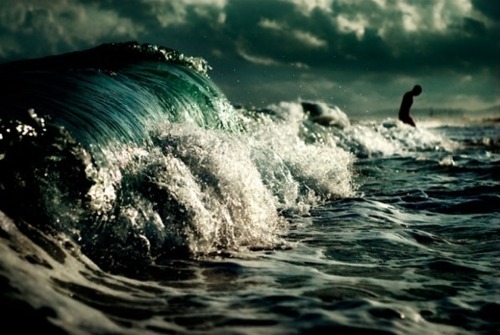 A close-up of waves crashing under stormy skies, with a distant figure providing scale to the white foam and teal-grey water breaking with force.