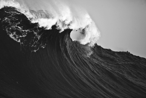 A monumental wave reaches its peak in black and white, white foam cascading from the crest in an almost architectural form that emphasizes the sculptural quality of moving water.