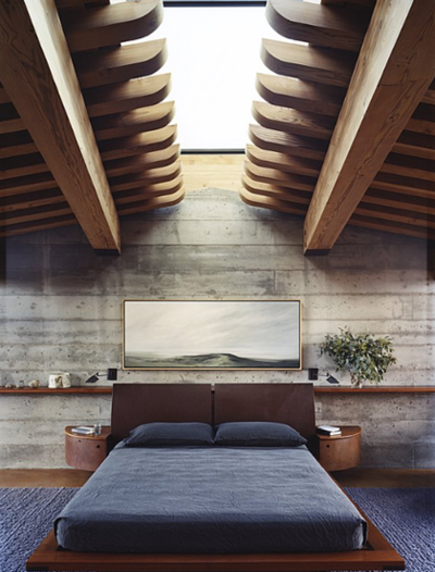 Dramatic wooden ceiling beams frame a skylight above raw concrete walls in a contemporary bedroom. The geometric shadows and minimal platform bed create a zen-like space balancing industrial materials with serene comfort.