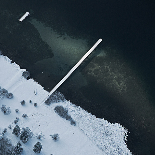 An aerial view reveals a white pier extending into dark teal-black water, while snow blankets the shoreline in organic curves. Scattered trees dot the frozen landscape, creating stark geometric contrast.