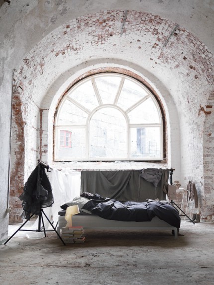A dramatic arched window illuminates a minimal bedroom in a converted stone structure with white-painted brick walls. The simple bed beneath the semi-circular window creates a monastic quality within the industrial bones.