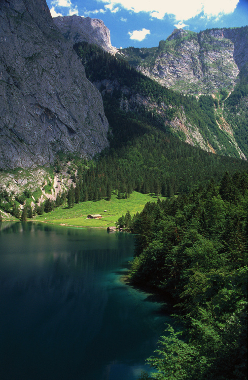 Towering limestone cliffs frame an emerald-green alpine lake, with dense coniferous forests cascading down the mountainsides. A small rustic structure on the distant shore provides scale to this dramatic valley landscape.