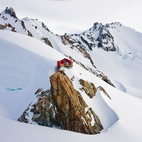 A red mountain hut perches on a jagged rock outcrop surrounded by pristine snow-covered peaks and glacial terrain. The small shelter creates powerful contrast between human presence and the austere high-altitude wilderness.