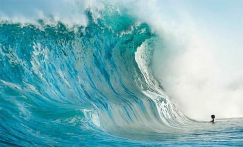 A magnificent turquoise wave crashes with tremendous force, its crystalline barrel creating a perfect tube while a distant surfer sits dwarfed by the ocean's power.