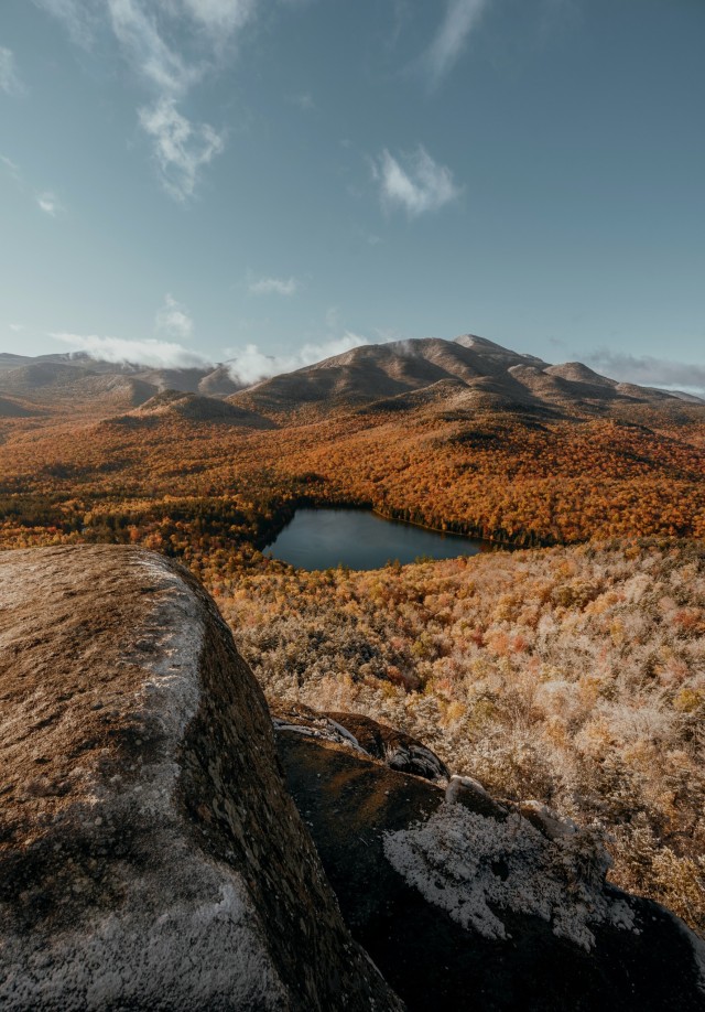 Photographed from a rocky outcrop, this Adirondack scene shows a mountain lake surrounded by golden-orange fall foliage beneath dramatic rolling peaks. The elevated viewpoint emphasizes the wilderness scale and seasonal transformation of the valley.