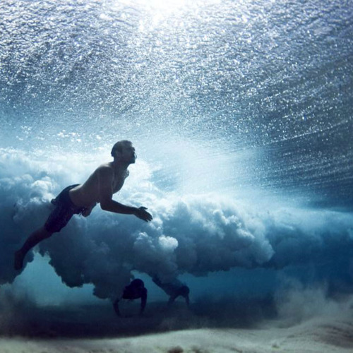 Photographed from below looking up, a silhouetted swimmer moves beneath turbulent waves, with churning white foam and bubbles creating a dynamic ceiling of breaking surf.