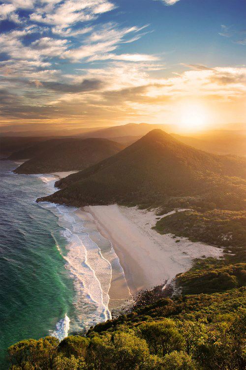 Aerial perspective reveals a secluded beach nestled between rolling coastal hills as the sun breaks through clouds at golden hour. Emerald water and pristine sand glow under warm light that illuminates both terrain and sea surface.