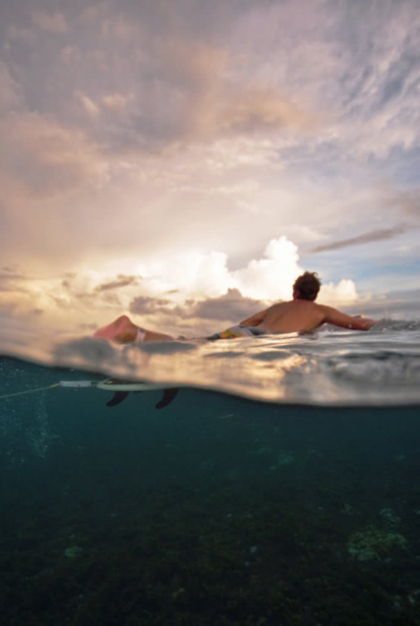 A split-level underwater photograph captures a figure floating on the surface during golden hour, with the illuminated sky above reflected in ripples while darker depths reveal the underwater world below.