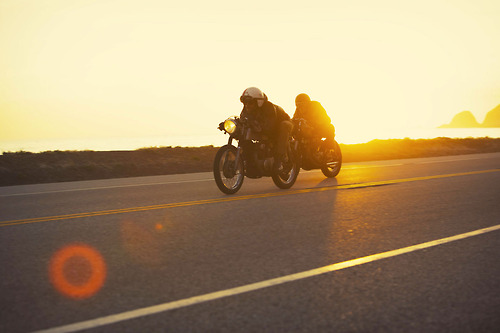 A vintage motorcycle with white-walled tires sits on weathered cobblestones against classic European architecture at golden hour. Dramatic lens flares and warm shadows across the textured stone surface create nostalgic, cinematic atmosphere.