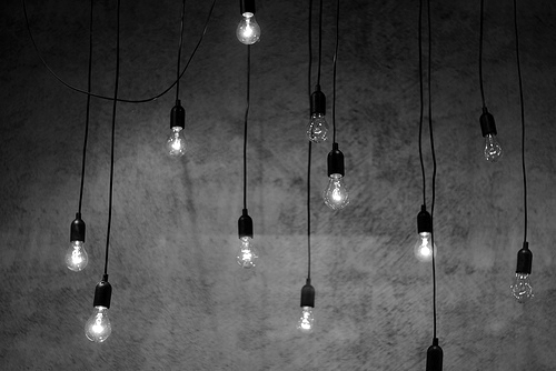 Multiple Edison bulbs hang at varying heights against a textured concrete wall in an artfully casual arrangement. The exposed pendant lights create intimate pools of warm illumination against the raw industrial surface.