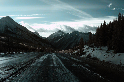 A winter highway ribbons through a mountain valley toward snow-capped peaks shrouded in clouds, with evergreen forests and snow patches flanking the dark asphalt. The overcast sky and empty road capture the raw solitude of mountain wilderness.