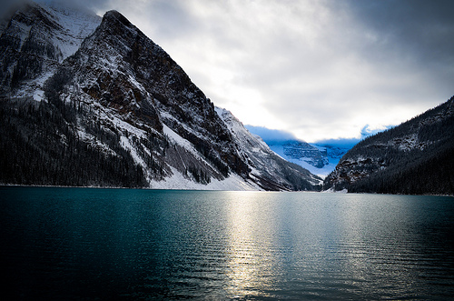Steep granite cliffs rise directly from the edge of a glacial lake, their snow-dusted surfaces reflected in the still water below. The overcast sky suggests changing weather in this pristine alpine setting.