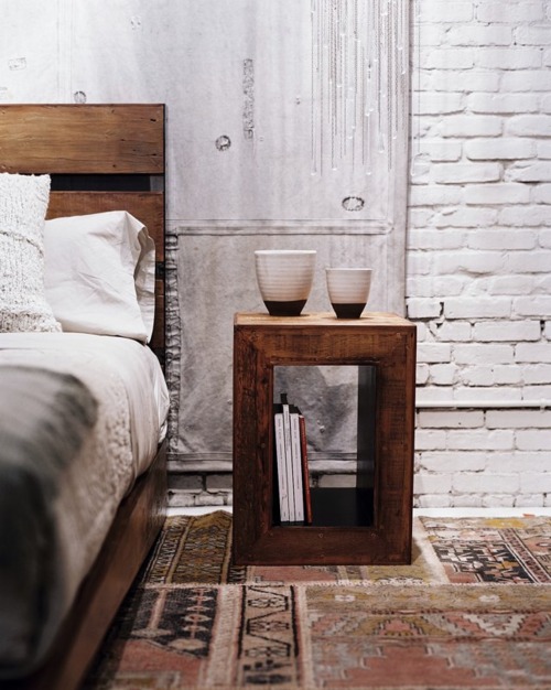 A reclaimed wood bed and cubic nightstand sit against weathered concrete walls and exposed white brick. Two ceramic vessels on the bedside table and vintage rugs below create warmth within the raw industrial bedroom.