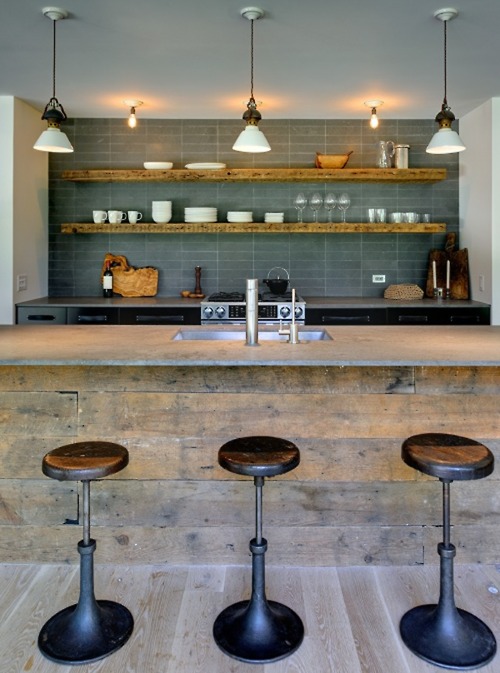 White-painted ceiling beams cross above a kitchen with dark brick walls and stainless steel appliances. A central island with galvanized metal stools anchors the space while open shelving displays white dishware against the moody backdrop.