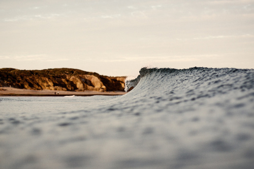 A translucent wave curls in the foreground while golden coastal cliffs rise in the background, capturing the moment where ocean energy meets weathered stone.
