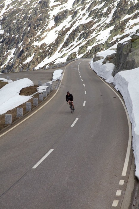 A lone cyclist navigates a winding mountain road between towering snow-covered peaks and deep snow banks. The pristine asphalt cuts through stark winter terrain marked by weathered stone barriers.