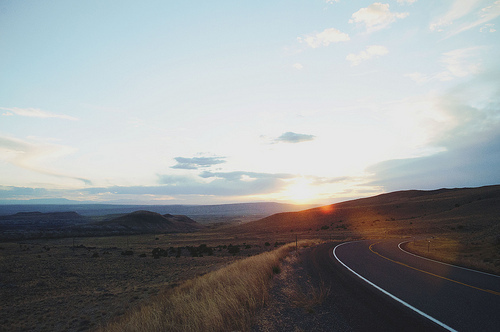 An empty highway curves through rolling desert hills at golden hour, the sun setting low on the horizon and casting warm light across sparse vegetation. The road stretches into the distance under a pale sky, creating a sense of infinite journey.
