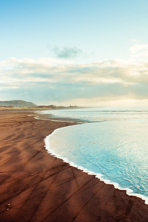 Dark volcanic sand curves along turquoise waters under a pastel sky, with natural tire-track patterns visible in the wet sand. The headlands fade into soft atmospheric haze in the distance.