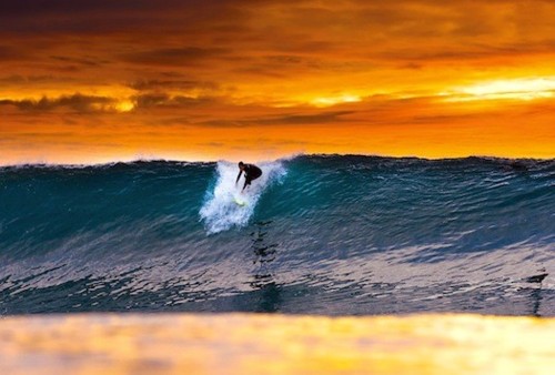A surfer carves through a turquoise wave at sunset, with fiery orange storm clouds behind and the golden light reflecting across the water's surface.