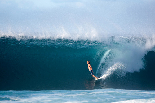 A surfer rides inside the barrel of a massive turquoise wave, with crystalline water forming a translucent tunnel and white foam cascading overhead against a moody sky.