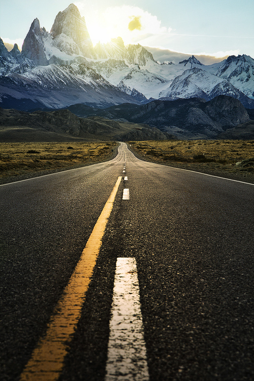 A straight asphalt road with yellow center lines and white dashed markings stretches toward dramatic snow-capped peaks, creating a powerful vanishing point composition. Golden grassland spreads across the middle ground beneath the towering jagged mountains.