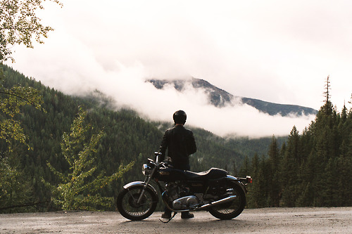 A motorcyclist sits contemplatively beside a vintage bike on a mountain road, silhouetted against mist-shrouded peaks and dense evergreen forests. Low-hanging clouds create an ethereal veil across the mountainous landscape, emphasizing solitude and adventure.