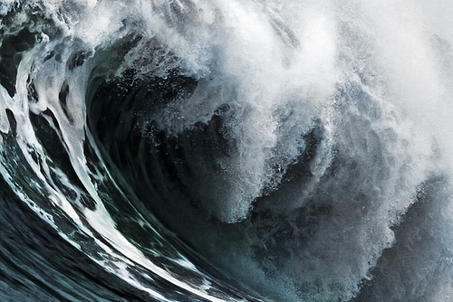 A wave breaks in a curved wall of deep blue-green water crowned with explosive white foam, freezing the raw energy and revealing the intricate textures within the barrel.