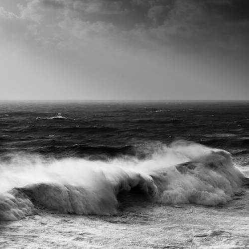 This black and white seascape emphasizes turbulent waves crashing in the foreground while storm clouds gather across the horizon, creating a study of natural forces in motion.