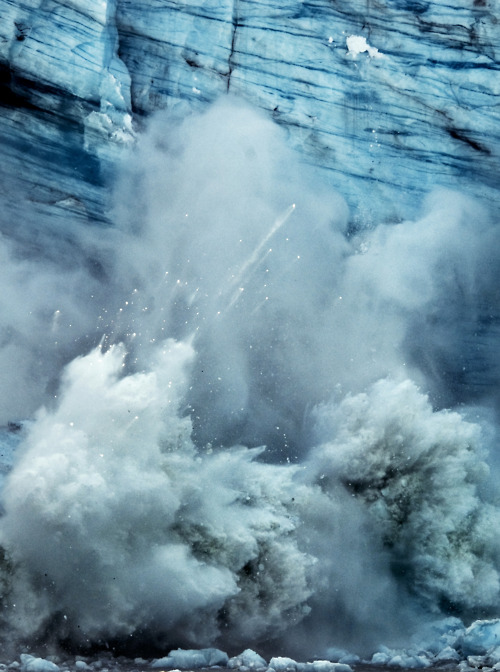 Churning white foam and spray erupt against layered blue ice formations in this study of elemental forces, showing the violent collision between water and glacial structures.