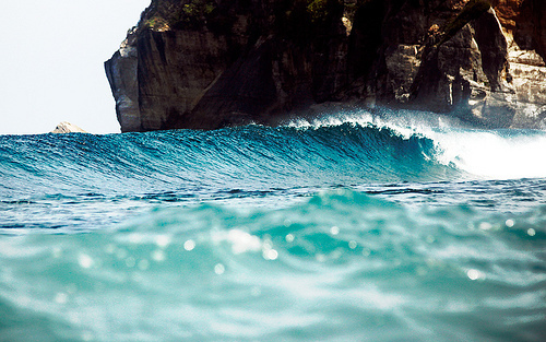 Shot from water level with crystalline droplets in the foreground, a turquoise wave breaks against a rocky cliff face, creating an immersive perspective directly in the ocean.