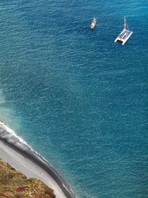 Two white sailboats anchor in a crescent bay where verdant hillside meets crystalline turquoise water. The aerial perspective captures the complete curve of the coastline and the boats' placement in the vast blue expanse.