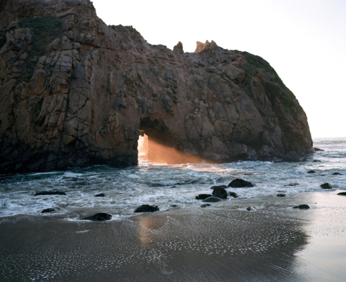 A natural rock arch frames golden light filtering through to a beach dotted with smooth stones. Gentle waves wash over the wet sand, creating reflections beneath the dramatic coastal formation at evening.