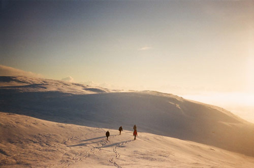 Three figures traverse unmarked snow during golden hour, their tracks cutting delicate lines across the mountainside. The warm amber sky contrasts with the pristine white slopes of the rolling peaks.