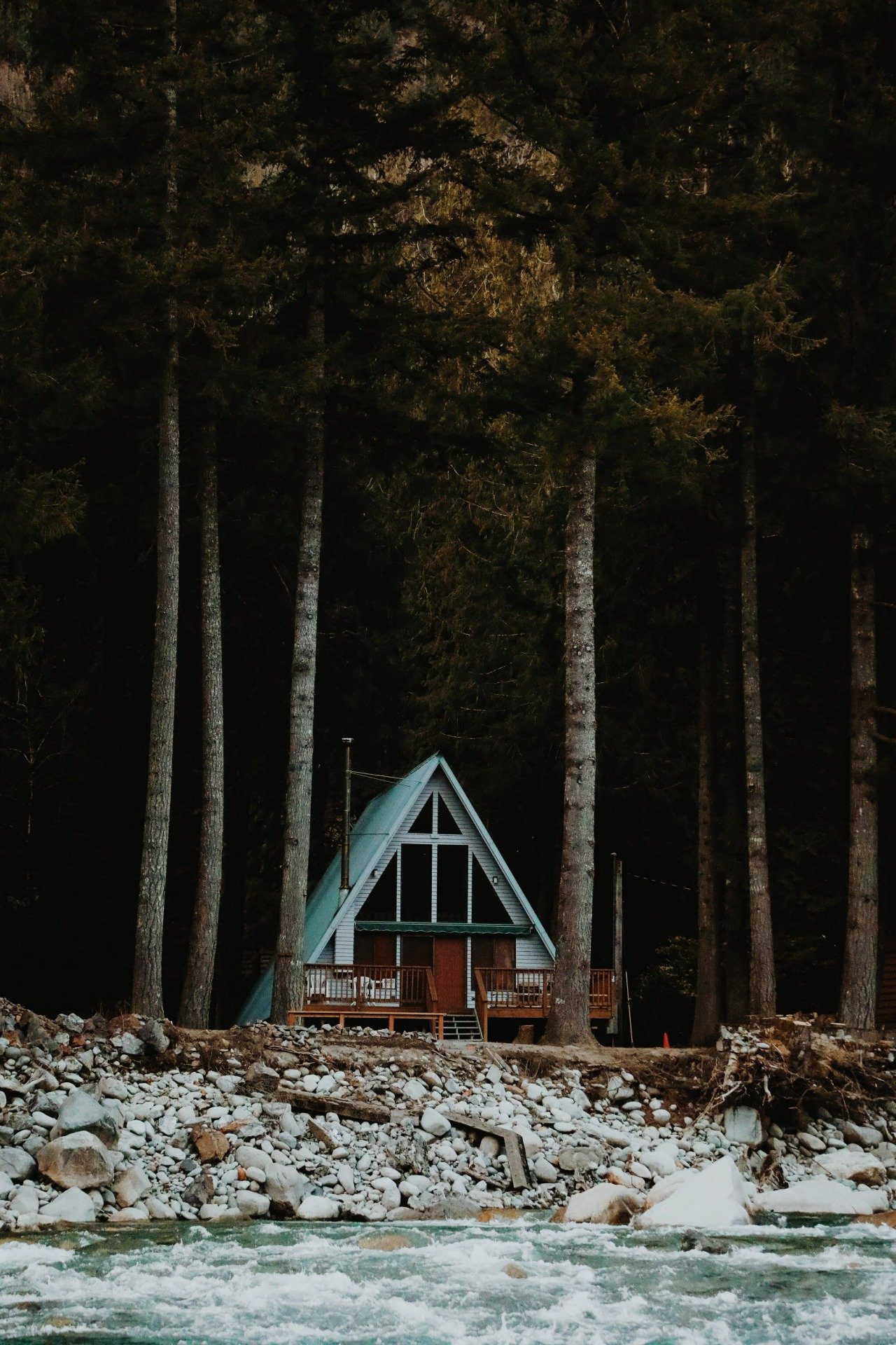 Teal A-Frame Cabin On Rocky Riverbank Dense Forest