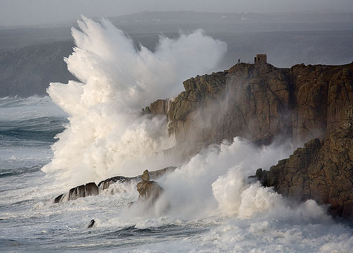 Massive waves crash against coastal cliffs with explosive force, sending towering plumes of white spray skyward while a solitary structure perches on the cliff edge.
